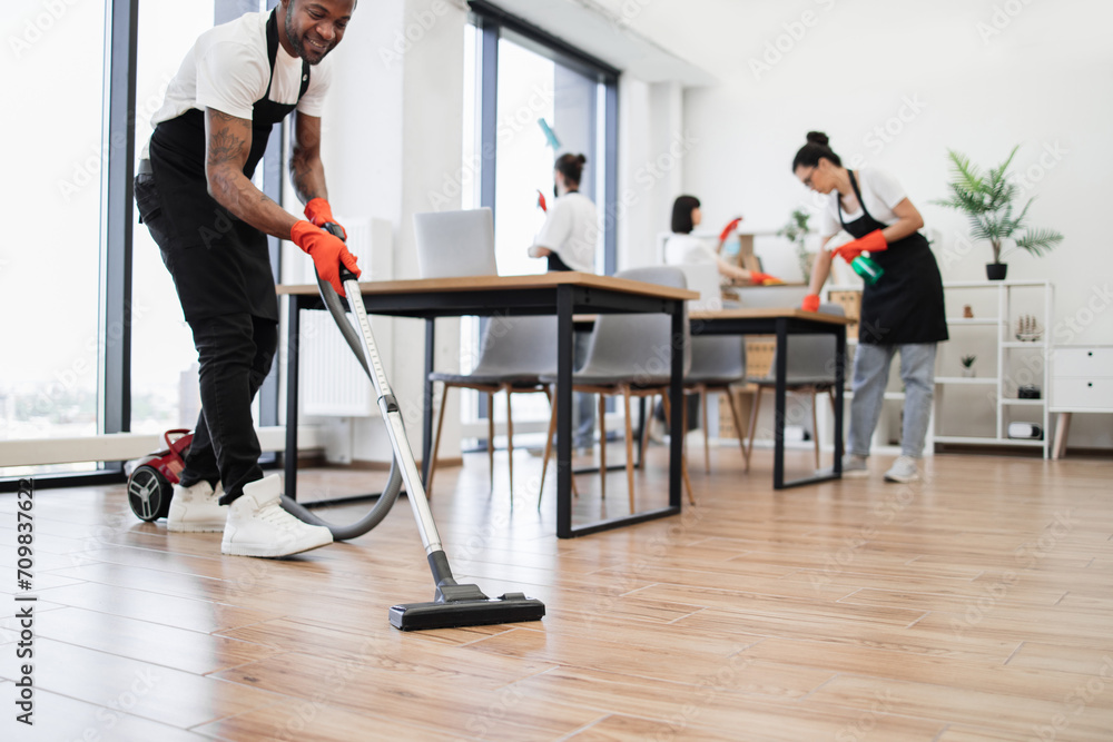 Cropped view of hands of African man holding vacuum cleaner. Team of young multicultural cleaners vacuums floor, wipes tables and gadgets, shelves, washes windows in spacious, bright, modern office.