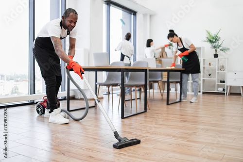 Side view of African man using vacuum cleaner to clean floor from dirt. Team of young multicultural cleaners wipes tables and gadgets, shelves, washes windows in spacious, bright, modern office.