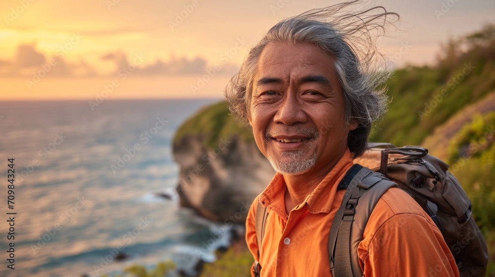 Active elderly Asian man with gray hair wearing sports wear for hiking and backpack is standing on the hill looking at the sunset over the ocean. He is happy and relaxed. Active elderly Asian man with gray hair wearing sports wear for hiking and backpack is standing on the hill looking at the sunset over the ocean. He is happy and relaxed.