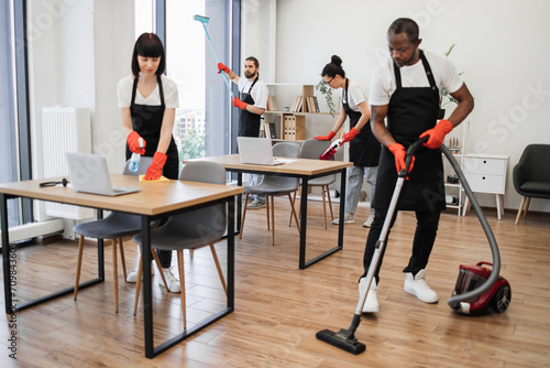 Focus on positive bearded Caucasian worker in a black apron and red rubber gloves cleaning panoramic window glass with mop, looking at camera in modern office with multiethnic co-workers.