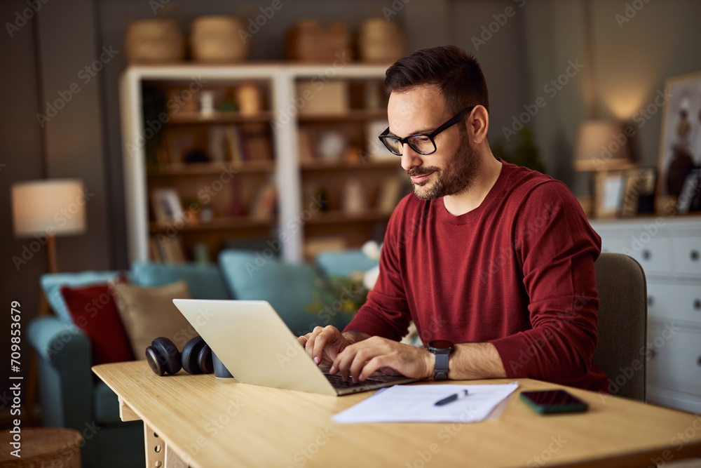 A focused male content creator typing on his laptop while working from home. - obrazy, fototapety, plakaty 