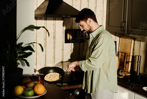 Side view of young man in grey cotton shirt holding hot frying pan while standing by electric stove and cooking omelet for breakfast photo