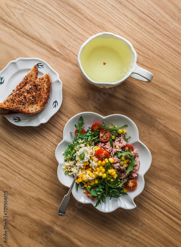 Green tea, crispy wholegrain toast and salad with canned tuna, boiled egg, arugula, cherry tomatoes and corn on a wooden background, top view