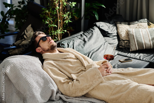 Restful guy in bathrobe and sunglasses holding glass of red wine in hand while relaxing on couch with pillows and daydreaming