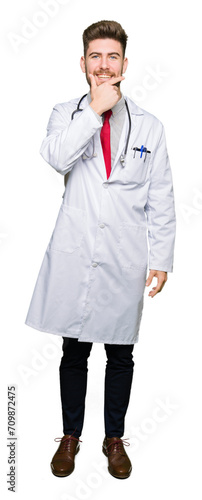 Young handsome doctor man wearing medical coat looking confident at the camera with smile with crossed arms and hand raised on chin. Thinking positive.