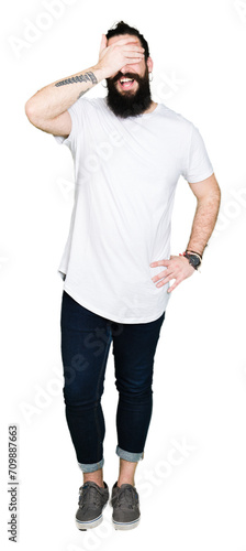 Young hipster man with long hair and beard wearing casual white t-shirt smiling and laughing with hand on face covering eyes for surprise. Blind concept.