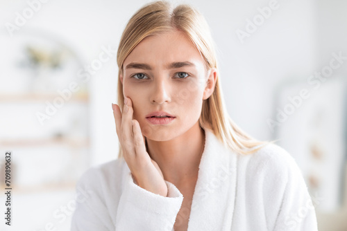 Worried blonde woman looking at camera touching cheek in bathroom