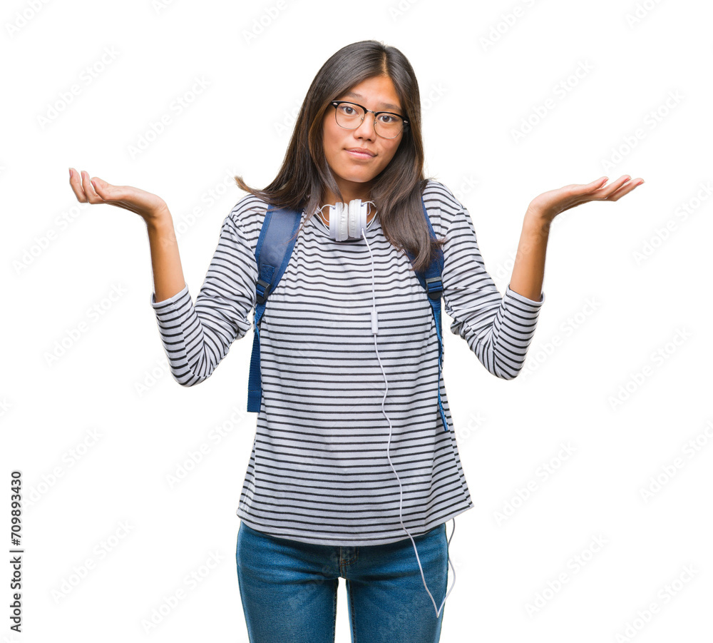 Young asian student woman wearing headphones and backpack over isolated background clueless and confused expression with arms and hands raised. Doubt concept. Young asian student woman wearing headphones and backpack over isolated background clueless and confused expression with arms and hands raised. Doubt concept.