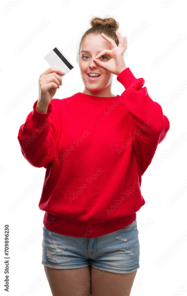 Young blonde woman holding credit card with happy face smiling doing ok sign with hand on eye looking through fingers Young blonde woman holding credit card with happy face smiling doing ok sign with hand on eye looking through fingers