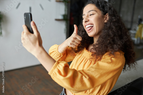 Side view of funny black girl of 20s recording short video or taking selfie on smartphone grimacing, making faces, sticking tongue and showing thumb up, having fun being in good mood