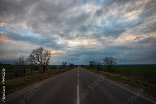 Empty evening highway among spring green meadows, clouds in the sky