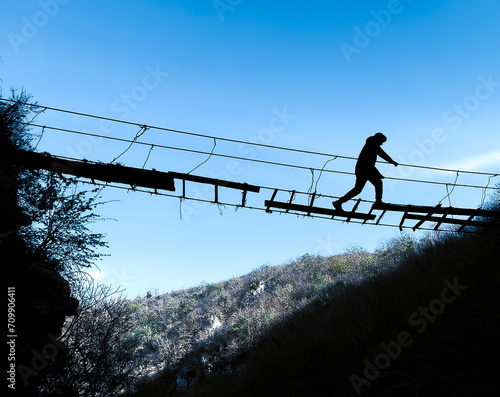 Silhouette of man walking over a suspension old bridge blue sky background