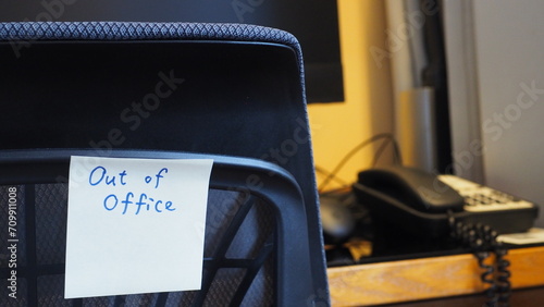 yellow sticky note with handwritten words "Out of office!" on back of chair, with office desk, computer and phone in the background 