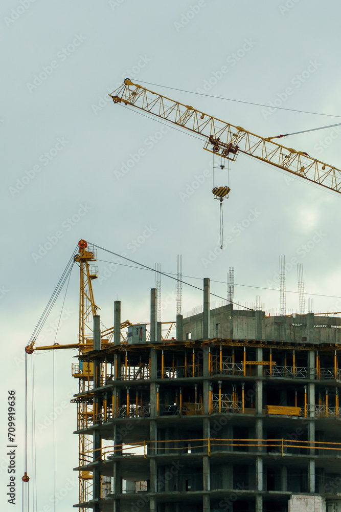 Silhouettes of working construction cranes against the background of an unfinished residential building. Multi-storey luxury apartment building on a construction site