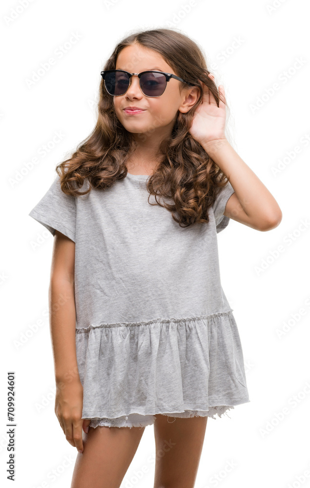 Brunette hispanic girl wearing sunglasses smiling with hand over ear listening an hearing to rumor or gossip. Deafness concept. Brunette hispanic girl wearing sunglasses smiling with hand over ear listening an hearing to rumor or gossip. Deafness concept.