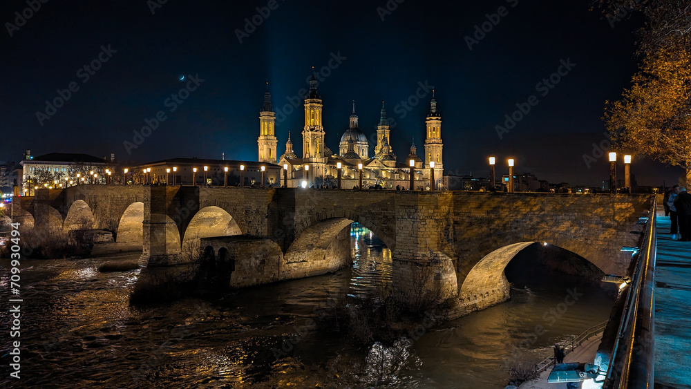 Moonlit Panorama: Zaragoza's Basilica del Pilar, Stone Bridge, and the Ebro River weave an enchanting tapestry in the tranquil glow of the evening moon.
