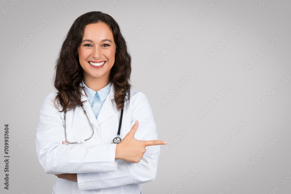 Cheerful lady doctor pointing finger aside at copy space advertising text of medical advertisement over grey background Cheerful lady doctor pointing finger aside at copy space advertising text of medical advertisement over grey background