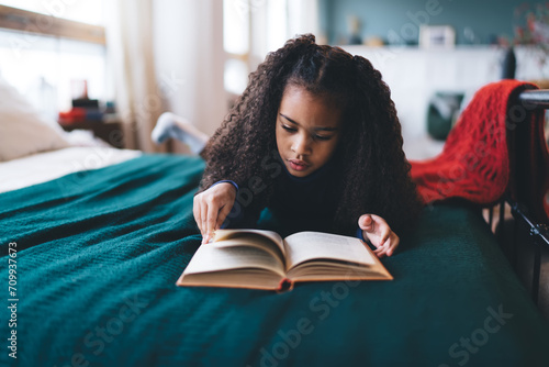 Focused black girl reading book while lying on bed at home