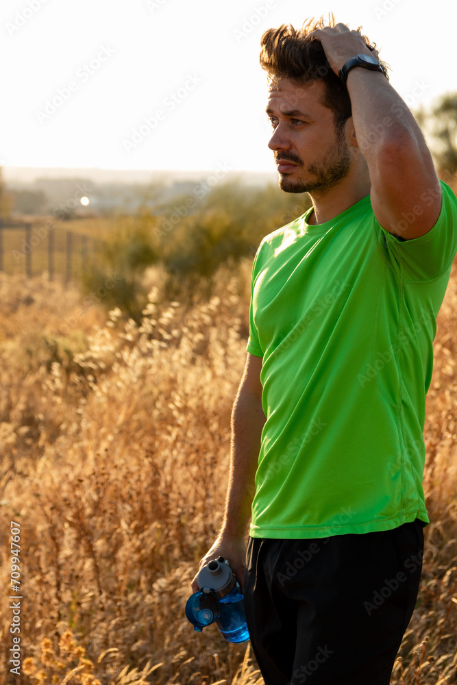 Fototapeta premium Young runner man with green t-shirt relaxing after sport training. Holding water bottle while doing fitness workout in summer
