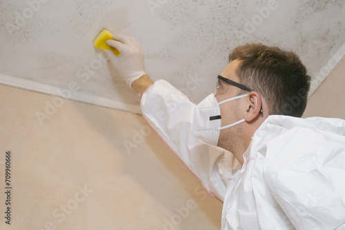 A cleaning service worker removes mold from a wall using a sprayer with mold remediation chemicals, mildew removers and a scraper. photo