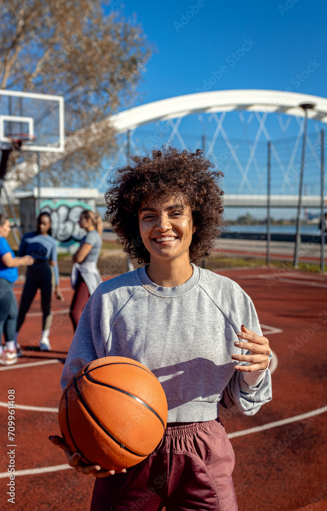 Portrait of young woman with her female friends on basketball court looking at camera and holding ball in her hands. Portrait of young woman with her female friends on basketball court looking at camera and holding ball in her hands.