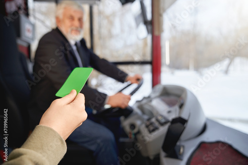Close up view of hand holding green payment card showing to driver while boarding in public transport. Crop of hand with paying ticket in front of blurred driver. Concept of transportation. photo