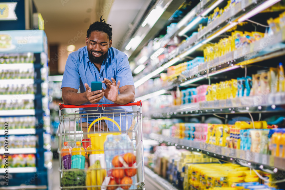 Black Male Buyer Shopping Groceries In Supermarket Taking Product From Shelf Standing With Shop Cart Indoors Using Phone.