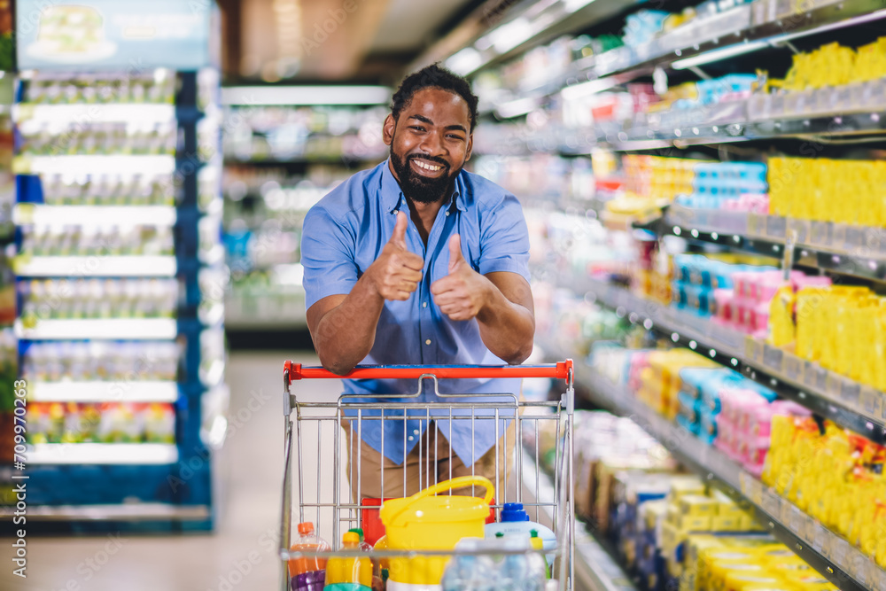 Black Male Buyer Shopping Groceries In Supermarket Taking Product From Shelf Standing With Shop Cart . Looking at the camera.Thumbs up.
