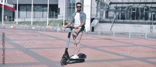 Freedom of movement. A handsome young adult man in stylish clothing and sunglasses confidently rides his electric scooter through the sunny city against the backdrop of modern buildings