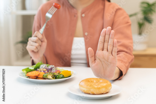 Diet concept  close up young woman  girl using hand push out  stop sweet donut  dessert or junk food on plate  choose green vegetables salad  eat low fat for good health. Female getting weight loss.