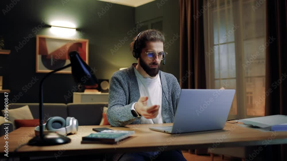 Male sitting at table in living room. Man conducting online meeting. Male working distantly. Man using laptop for video call. Male wearing glasses and headphones. Man typing at keyboard. Male sitting at table in living room. Man conducting online meeting. Male working distantly. Man using laptop for video call. Male wearing glasses and headphones. Man typing at keyboard.