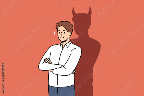 Confident businessman with bad thoughts stands with arms crossed near shadow with devil horns. Bad male office worker with treacherous ideas, wants to achieve goal in unethical way photo