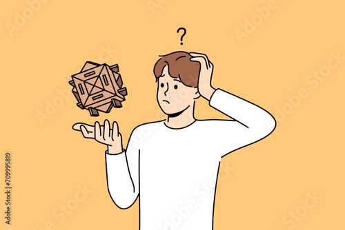Confused man with wooden puzzle scratches head, wondering how to solve difficult problem. Thoughtful guy with puzzle needs help to find solution due to lack of necessary qualifications