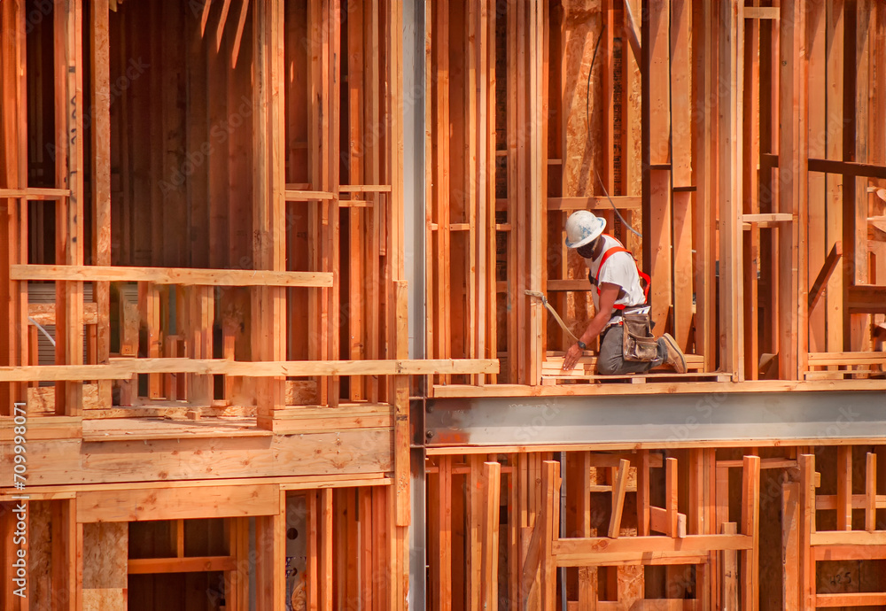 Horizontal image of a construction worker kneeling on wood framing wearing a tool belt, hard hat and safety harness. The shot is in afternoon light