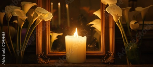 Conceptual image showing a large white candle illuminating arum lilies, with reflection on glass in a wooden frame.