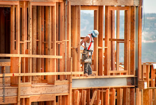 Horizontal image of a construction worker standing on wood framing wearing a tool belt, hard hat and safety harness. The shot is in afternoon light