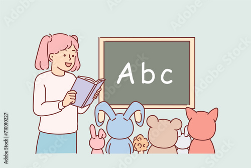 Little girl plays school teacher and reads book to toys sitting near blackboard with alphabet