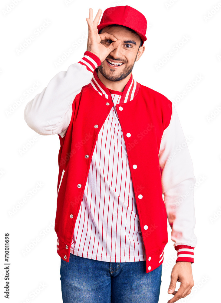 Young handsome man wearing baseball jacket and cap smiling happy doing ok sign with hand on eye looking through fingers Young handsome man wearing baseball jacket and cap smiling happy doing ok sign with hand on eye looking through fingers
