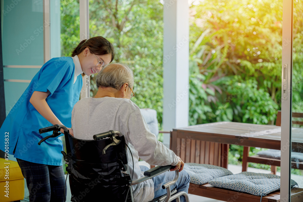 Fototapeta premium Young Asian nurse is supporting elderly man sitting in wheelchair walking to looking out window on garden and breathe fresh air, pension retirement center, senior man at home, healthcare concept