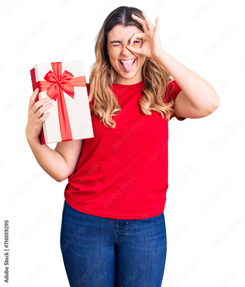 Young caucasian woman holding gift smiling happy doing ok sign with hand on eye looking through fingers Young caucasian woman holding gift smiling happy doing ok sign with hand on eye looking through fingers