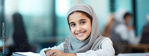 Smiling Muslim Arabic girl sitting at school desk in modern classroom. Back to school. Student with hijab. Islamic banner  photo