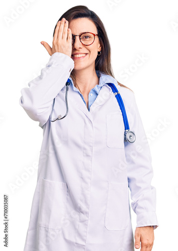 Young beautiful woman wearing doctor stethoscope and glasses covering one eye with hand, confident smile on face and surprise emotion.