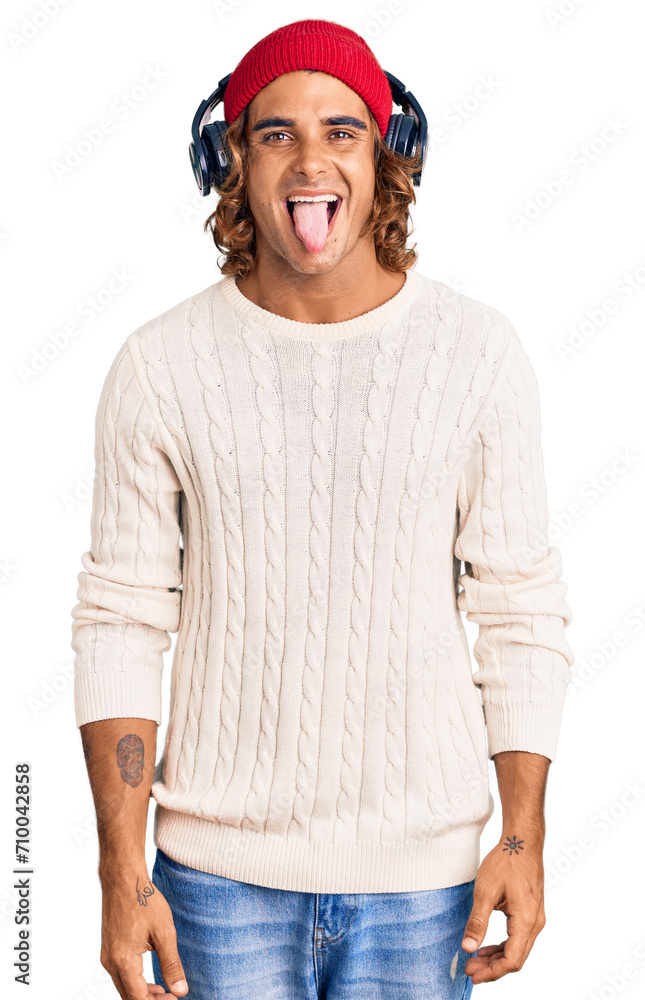 Fototapeta premium Young hispanic man listening to music using headphones sticking tongue out happy with funny expression. emotion concept.