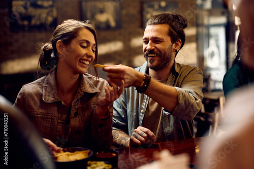Young happy man feeding his girlfriend while eating in pub.