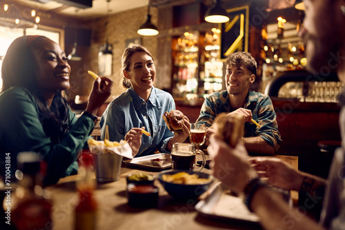 Multiracial group of happy friends eating burgers while drinking beer in pub.