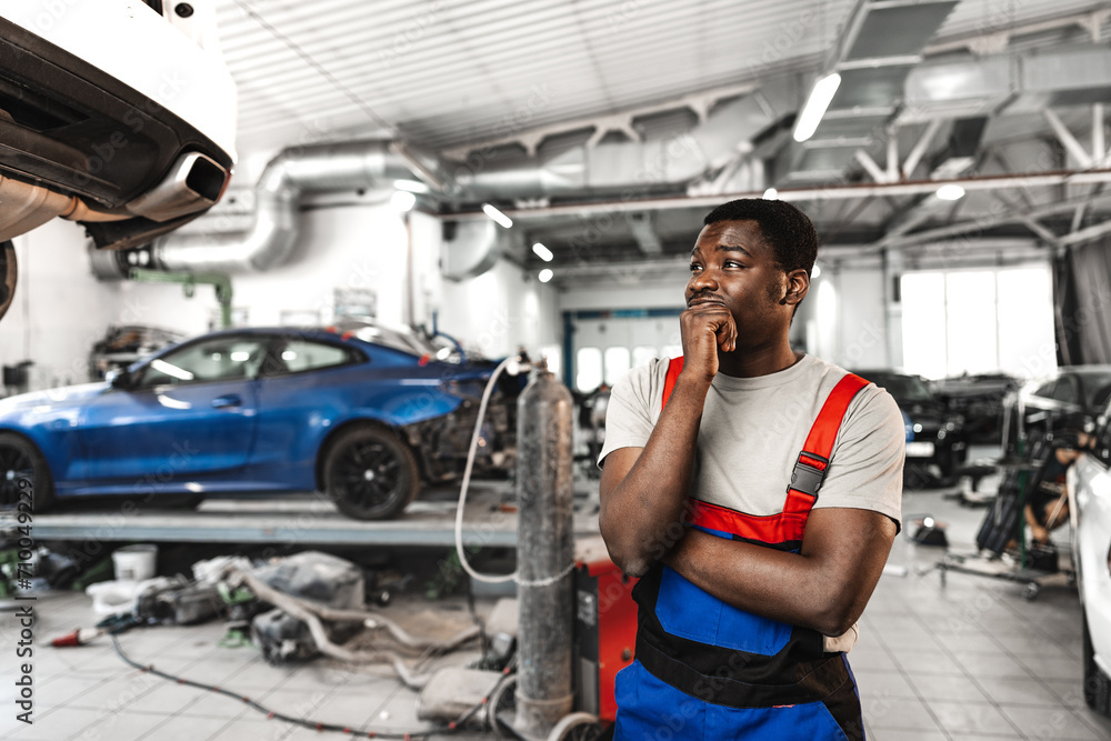 Pensive young African mechanic in car service looking at car thinking of how to repair it - obrazy, fototapety, plakaty 