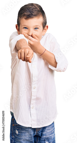 Cute blond kid wearing elegant shirt laughing at you, pointing finger to the camera with hand over mouth, shame expression