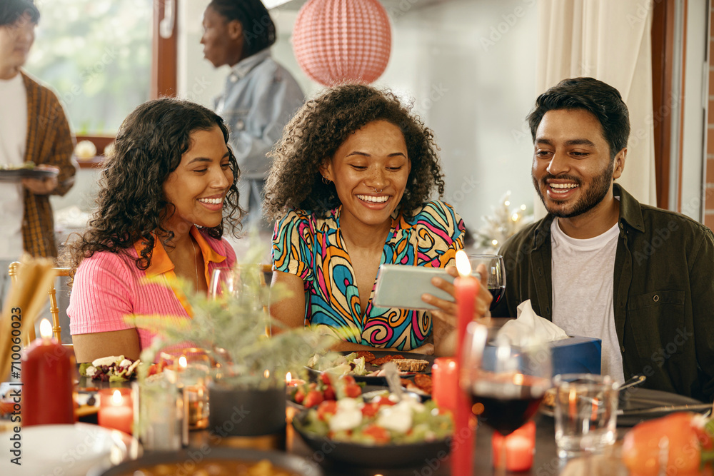 Smiling group of friends sitting around festive dinner table at home and looking at mobile phone Smiling group of friends sitting around festive dinner table at home and looking at mobile phone