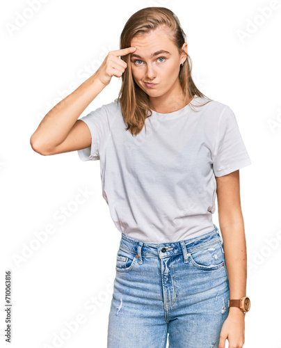 Beautiful young blonde woman wearing casual white t shirt pointing unhappy to pimple on forehead, ugly infection of blackhead. acne and skin problem