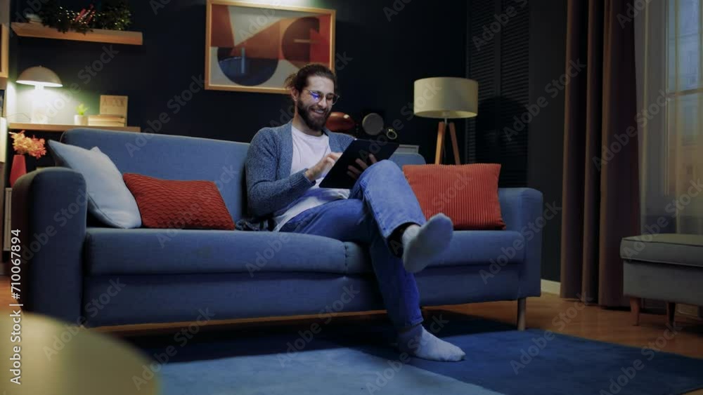 Cheerful man relaxing at home. Male sitting at sofa with crossed legs. Man holding tablet and laughing. Male wearing glasses and looking at tablet. Man having spare time at home. Staying at home. Cheerful man relaxing at home. Male sitting at sofa with crossed legs. Man holding tablet and laughing. Male wearing glasses and looking at tablet. Man having spare time at home. Staying at home.